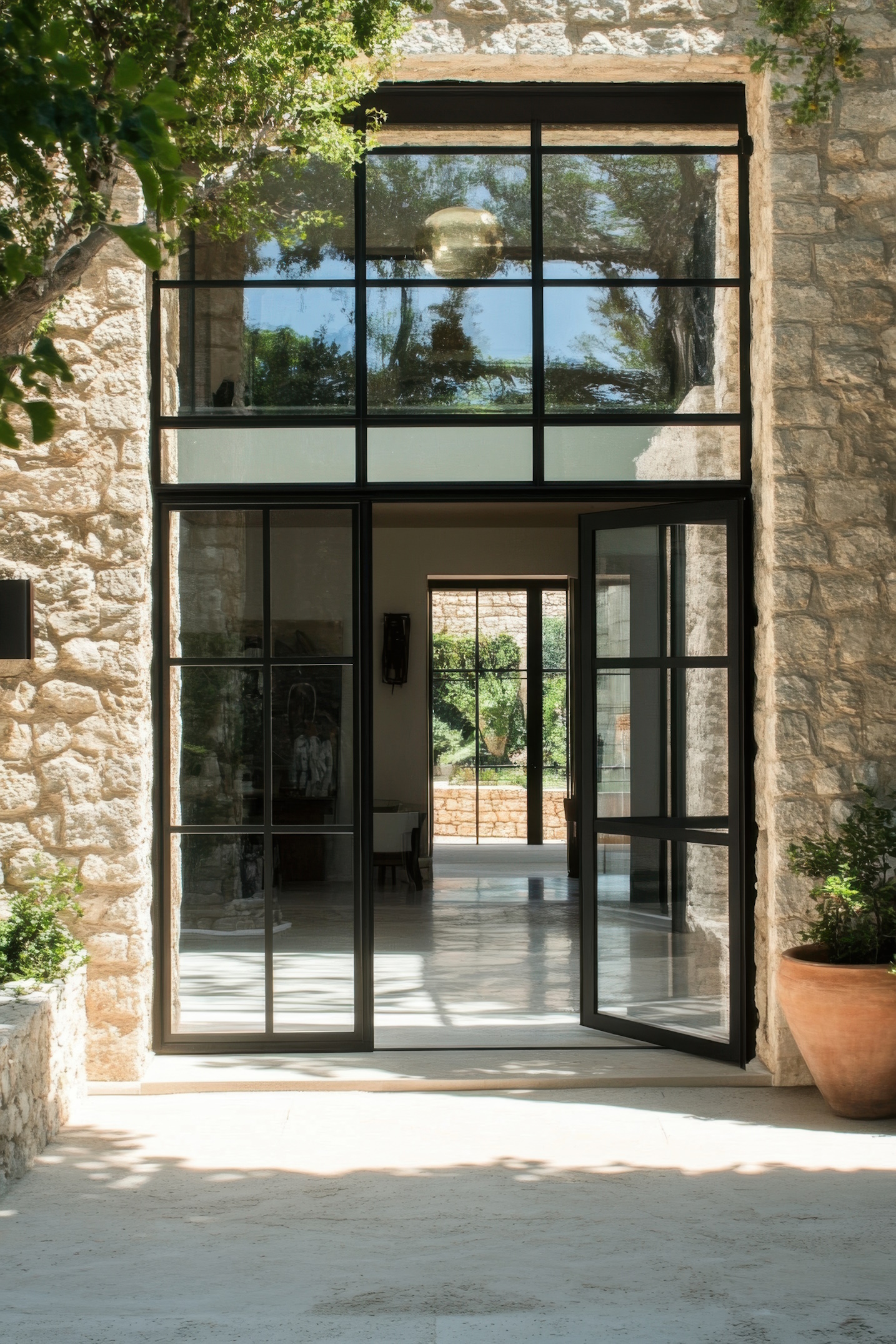 This modern stone house features a spacious entrance with large black-framed windows that allow natural light to flood the interior. Lush greenery enhances the welcoming atmosphere.